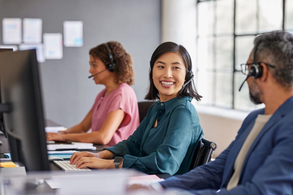 A diverse customer support team wearing headsets and working at their computers in an office. Illustrates VoIP-powered customer service in action.