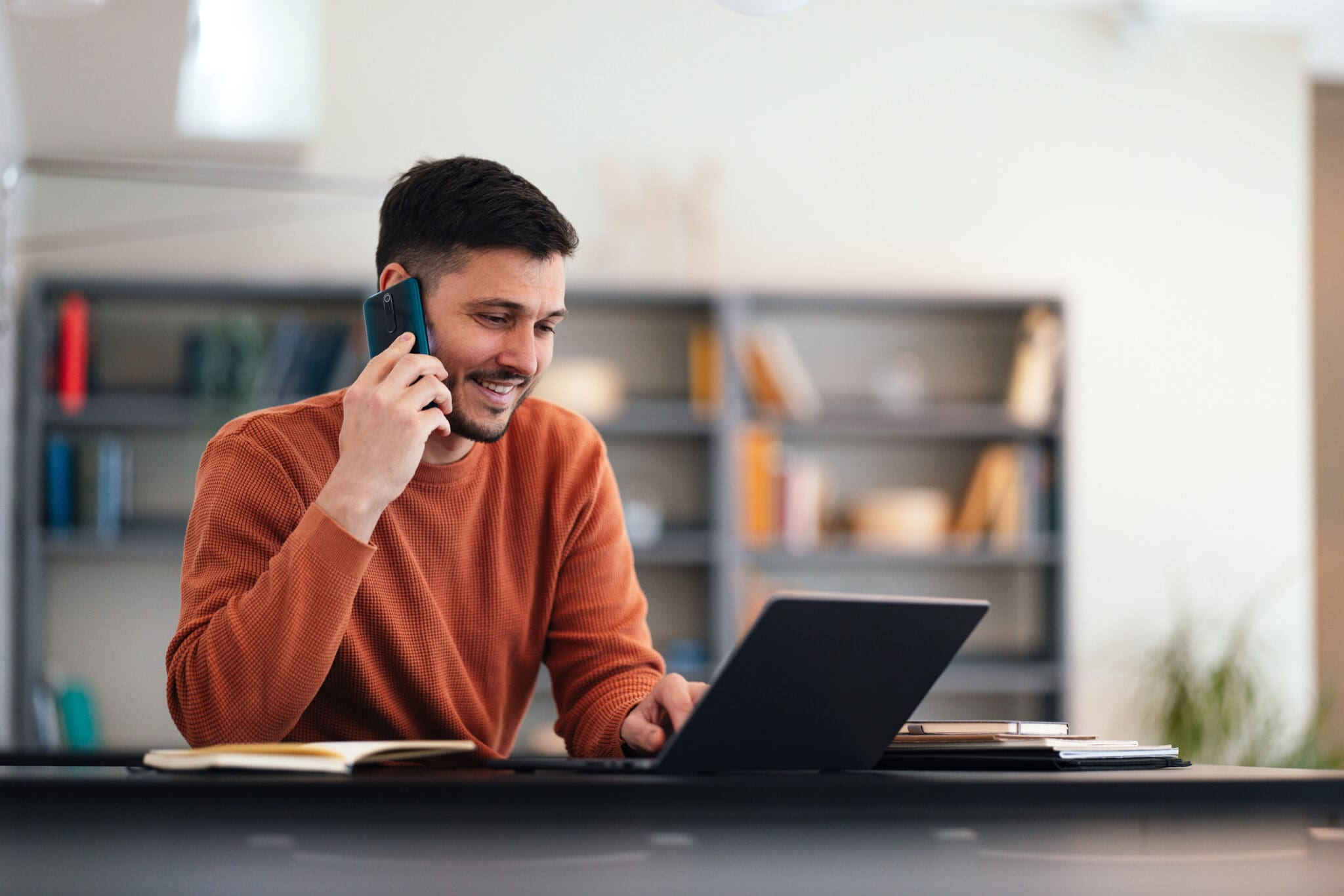 A man in a brown sweater talking on a VoIP phone while working on his laptop. Represents seamless VoIP communication for business efficiency.