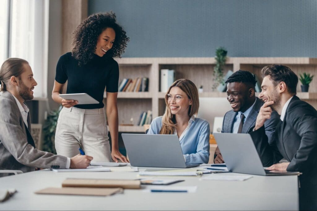 A diverse group of business professionals in a modern office, smiling and collaborating while using laptops and tablets, representing secure VoIP communication.