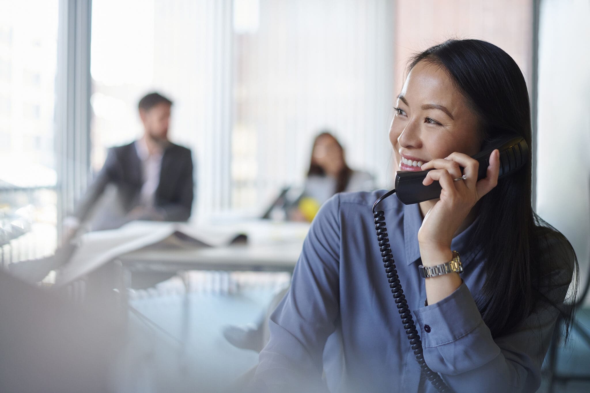 A professional woman in a modern office smiling while using a VoIP phone, with colleagues working in the background.