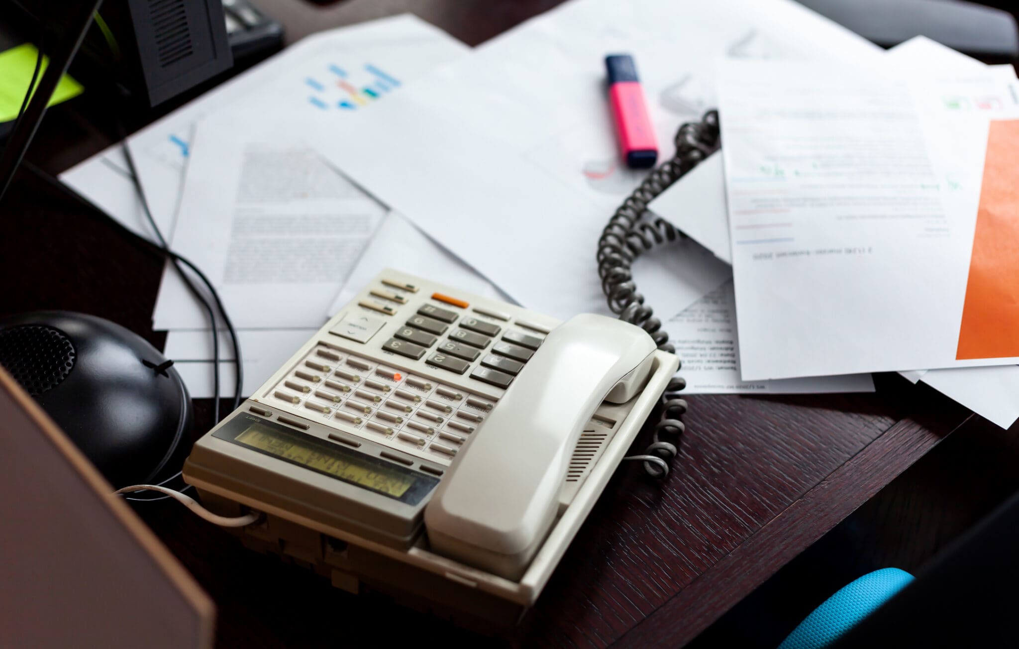 Cluttered office desk with a traditional landline phone, paperwork and cords, illustrating outdated communication technology.