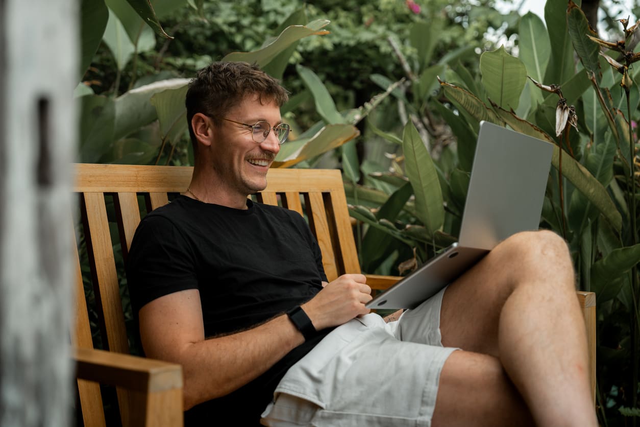 A remote worker sitting on a bench outdoors, surrounded by greenery, using a laptop and smiling.