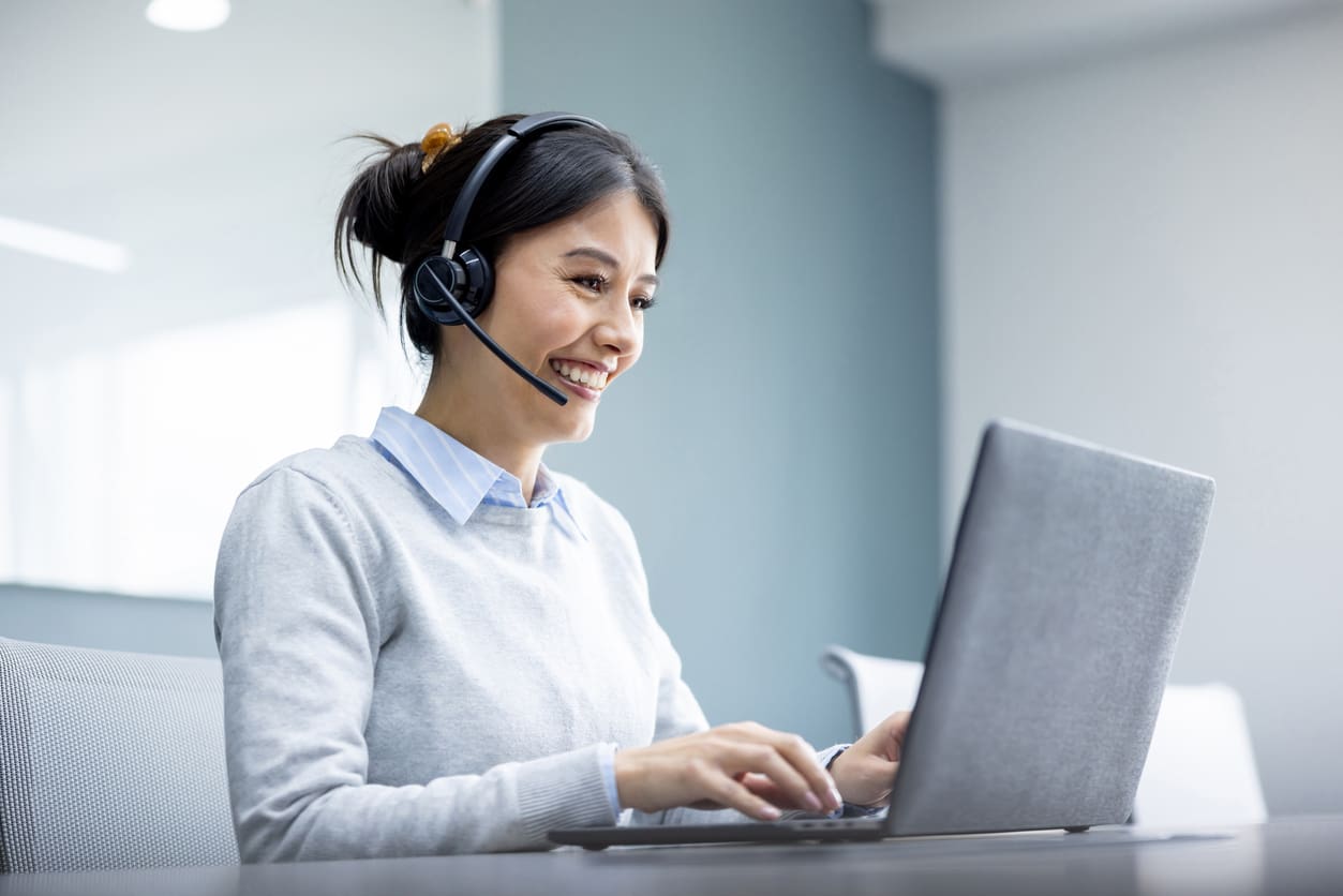 Smiling woman in a headset using a laptop for a VoIP call in a modern office.