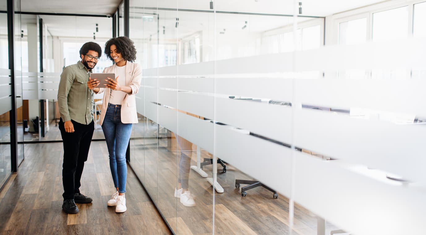Two professionals review information on a tablet in a bright office hallway, symbolizing collaboration and modern communication, powered by a VoiP system.