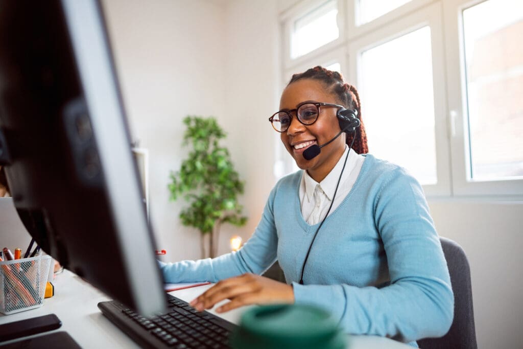 A smiling businesswoman wearing a headset, working at a computer in a bright home office, representing remote VoIP phone communication.