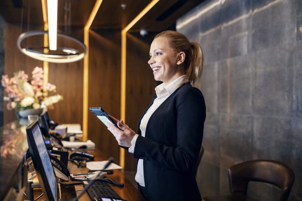 Smiling hotel front desk receptionist communicating via a tablet connected to a VoIP phone at the check-in counter.