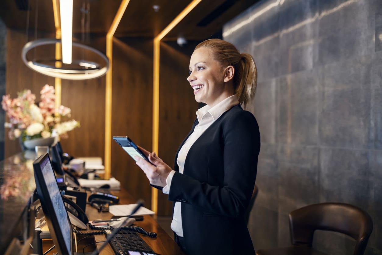 Smiling hotel front desk receptionist communicating via a tablet connected to a VoIP phone at the check-in counter.
